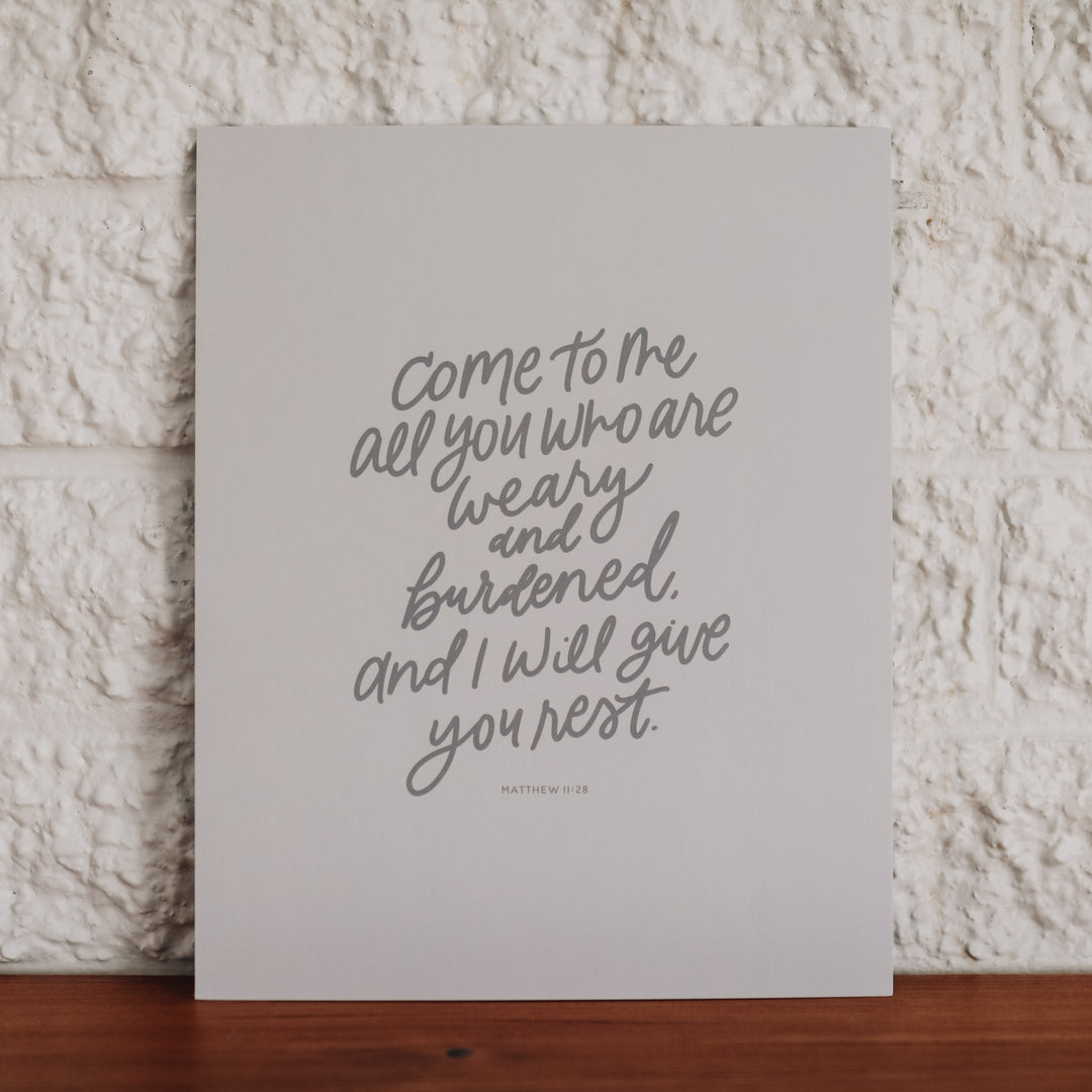 Print of Matthew 11:28 in a gray script next to a white brick wall. 